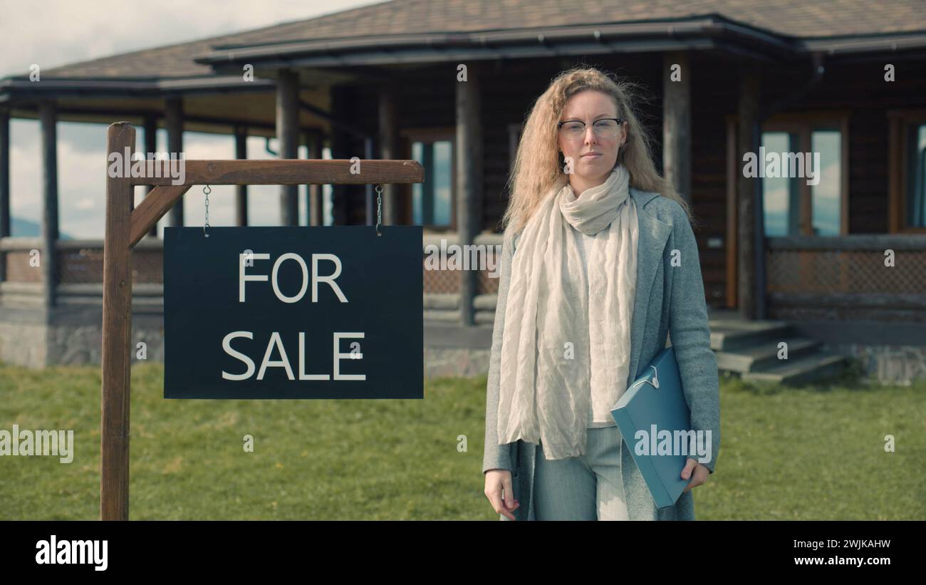 Female real estate agent stand near for sale sign and looks at camera. Realtor waits for