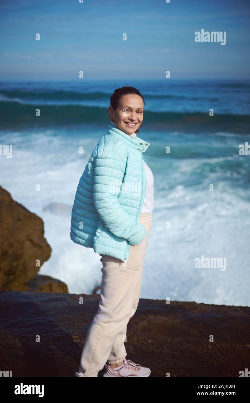 Happy smiling woman standing by ocean on the rocky cliff, looking at camera, enjoying the view of beautiful waves making white foam while crushing on Stock Photo