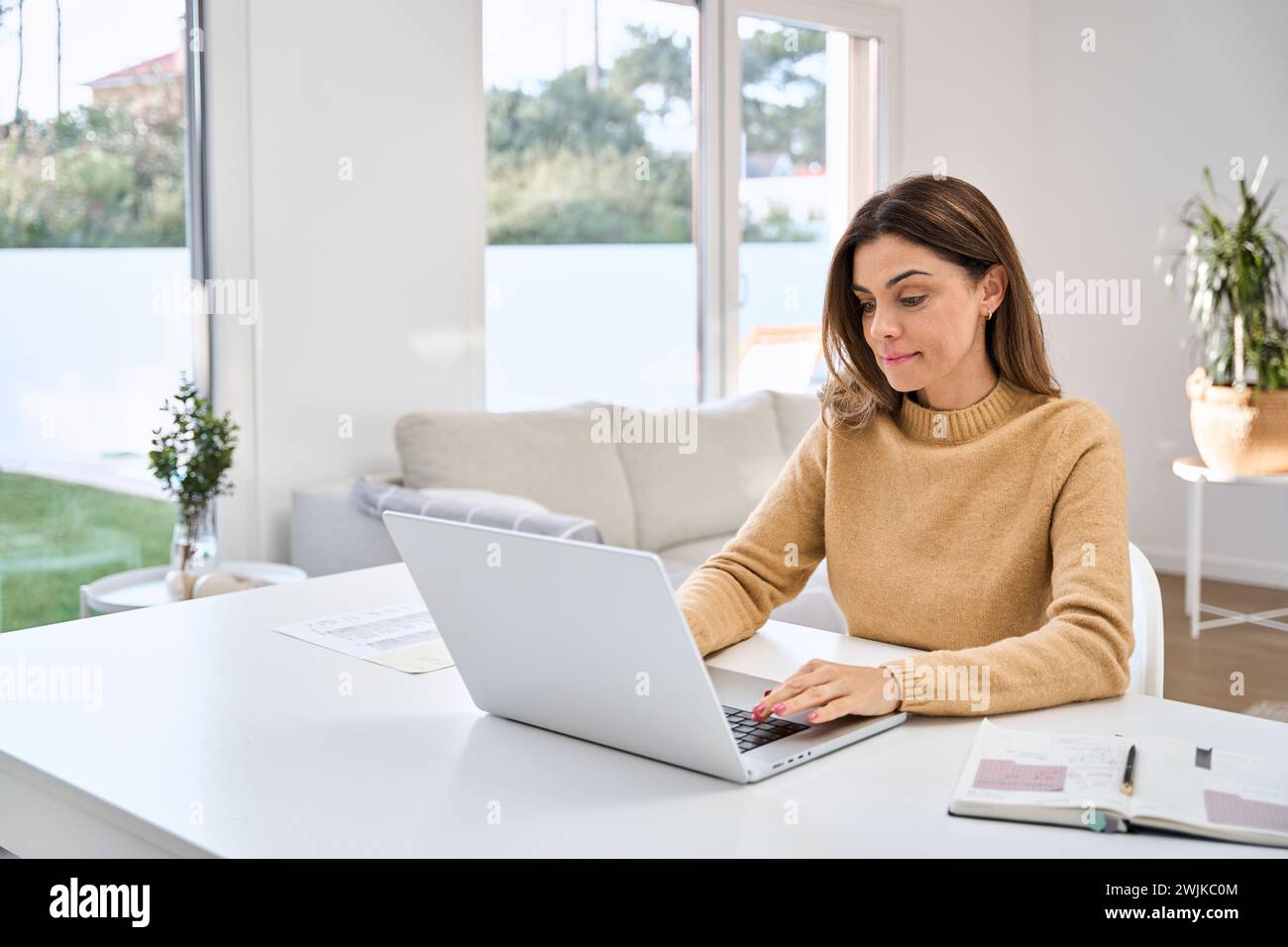 Smiling mature woman using laptop computer sitting at table in living room. Stock Photo
