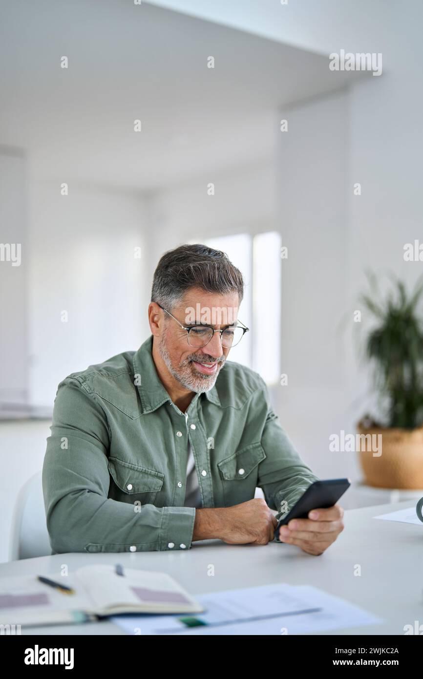 Happy middle aged man using cell phone sitting at home kitchen table. Stock Photo