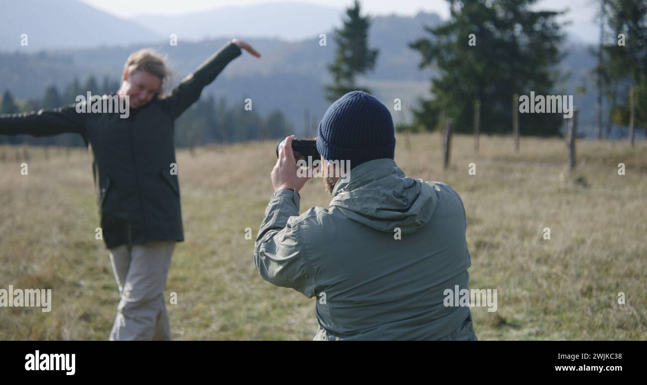 Caucasian man takes pictures of his wife on hilltop against scenic nature view using professional camera. Couple of travelers, tourist family during hiking trip or trek in mountains. Active leisure. Stock Photo