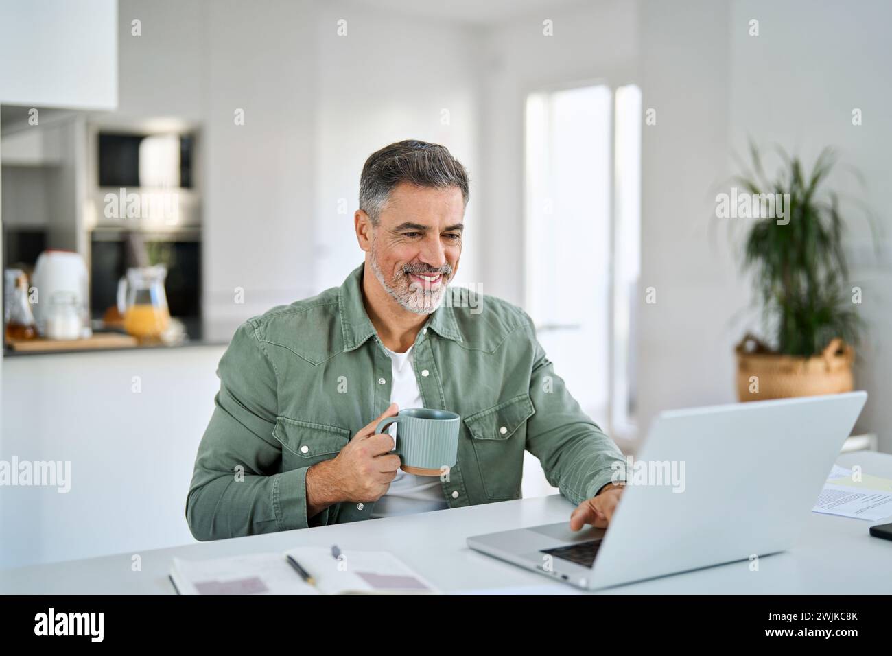 Happy older middle aged man drinking coffee using laptop at home table. Stock Photo