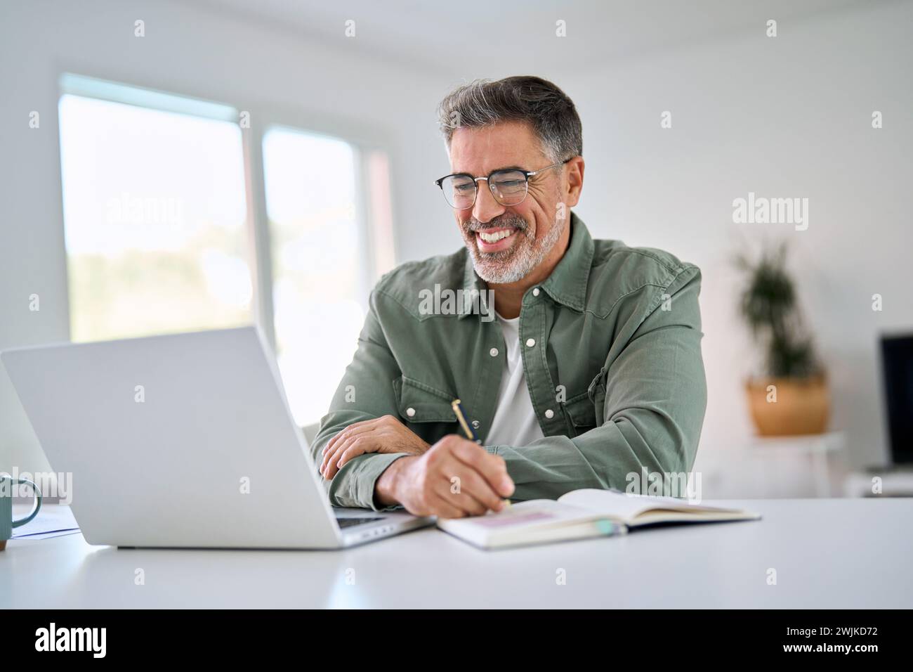 Happy middle aged man using laptop computer writing notes sitting at home table. Stock Photo