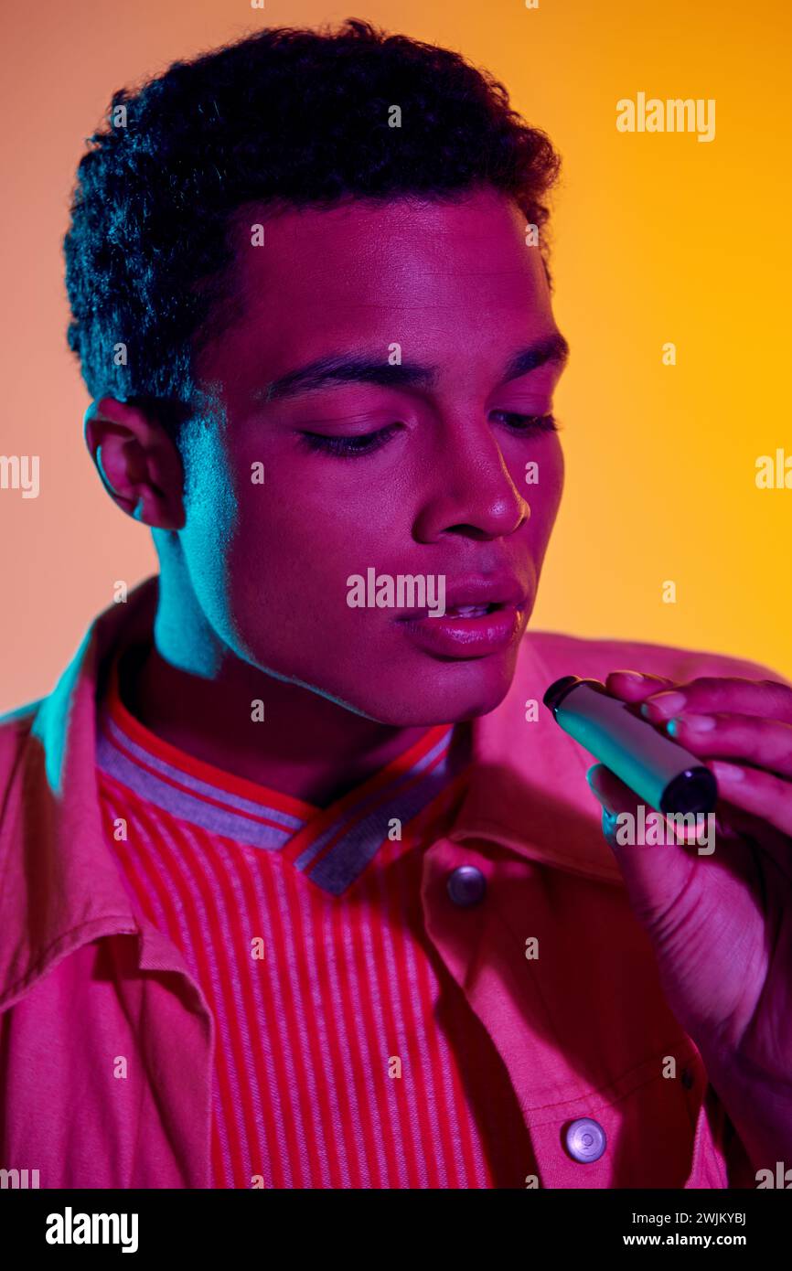 african american man vaping with electronic cigarette in hand, orange backdrop with lighting Stock Photo