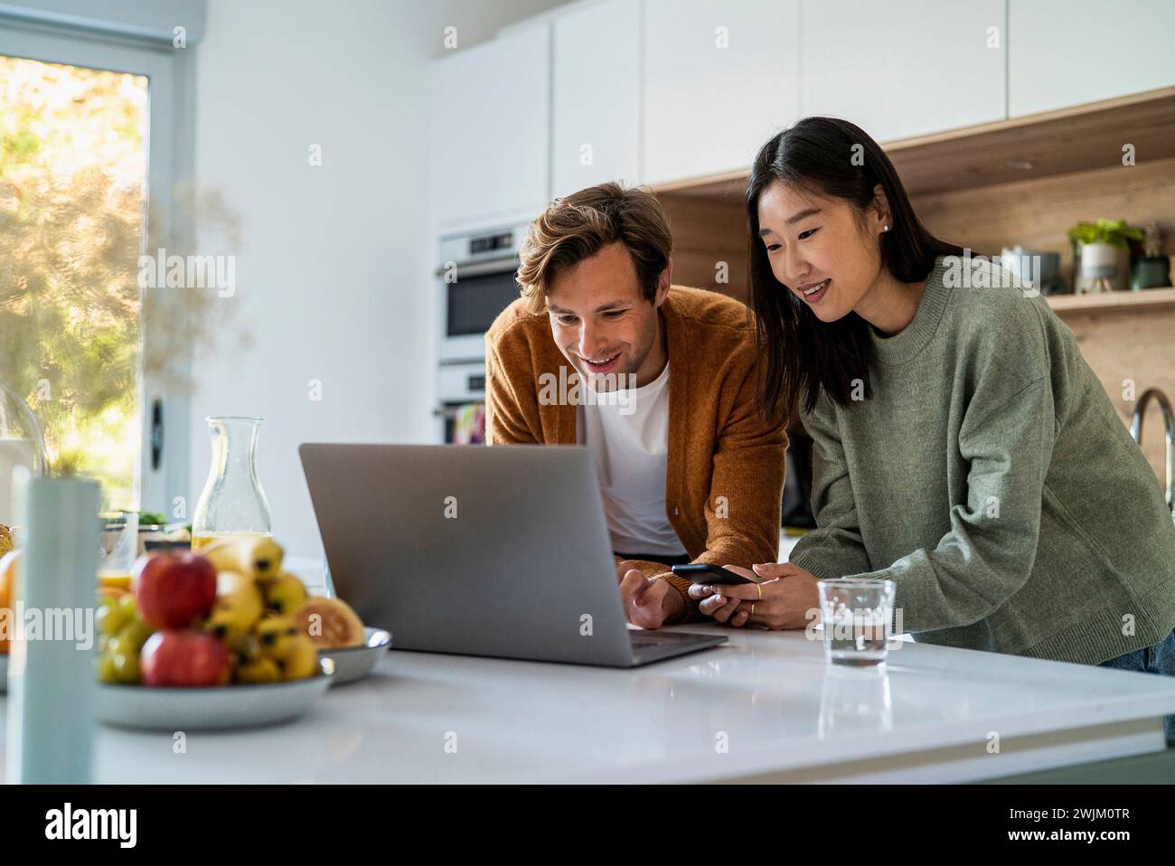 Adult couple shopping online on laptop and smart phone at kitchen counter Stock Photo