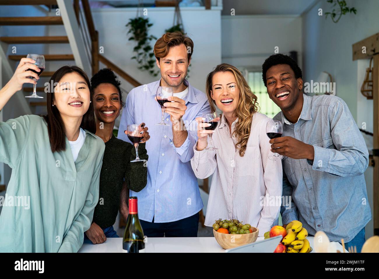 Group of friends looking at the camera toasting with wineglasses standing in kitchen Stock Photo