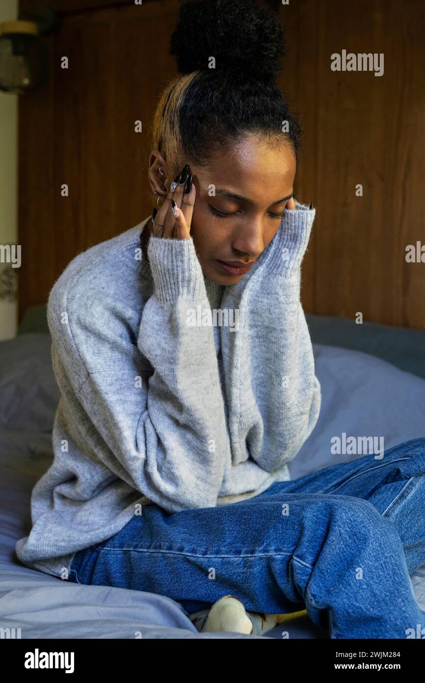 Stressed young adult woman touching her head while sitting on bed Stock Photo