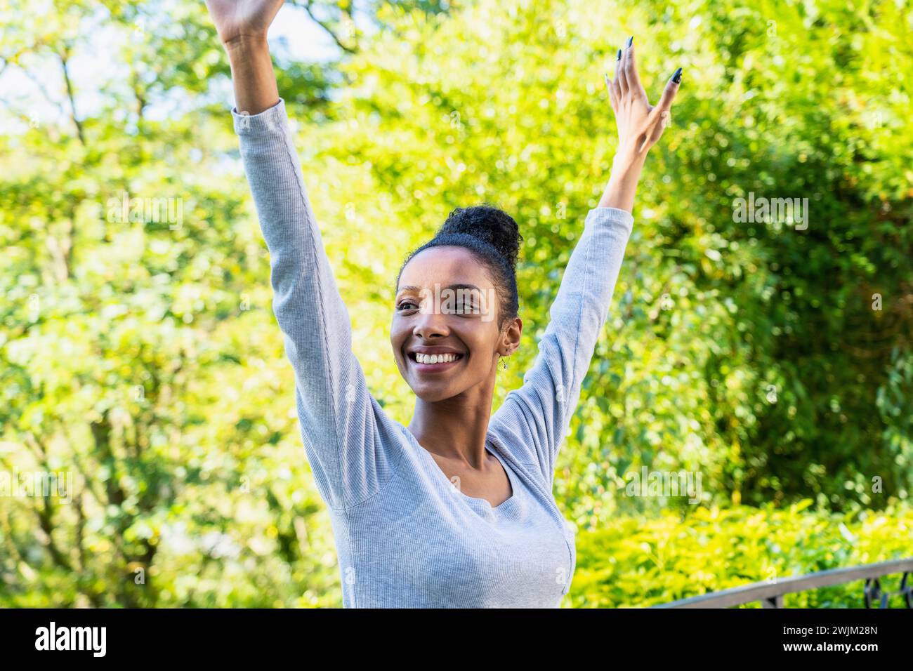 Cheerful young adult woman standing outdoors with arms raised Stock Photo