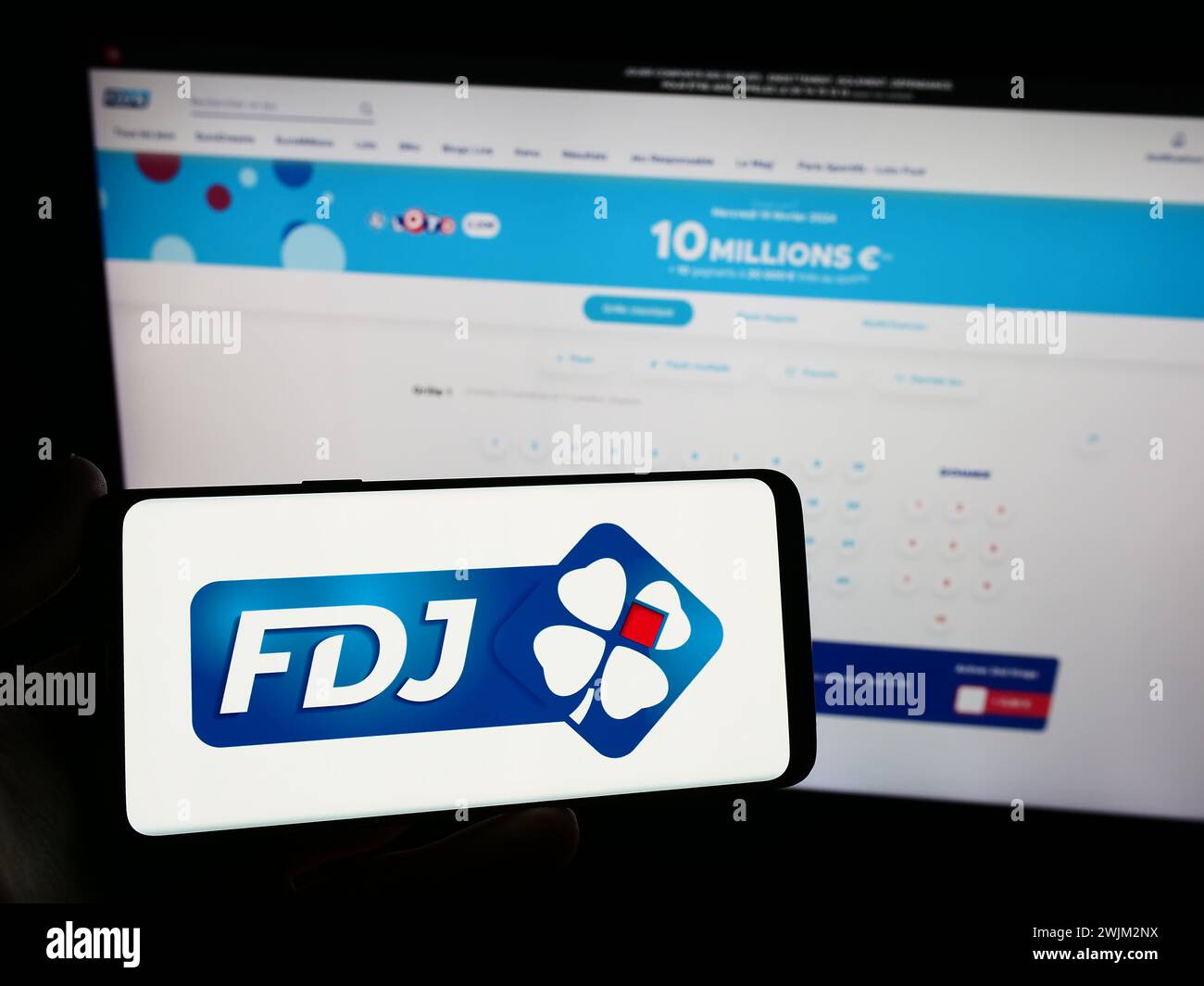 Person holding mobile phone with logo of French lottery company Francaise des Jeux (FDJ) in front of business web page. Focus on phone display. Stock Photo