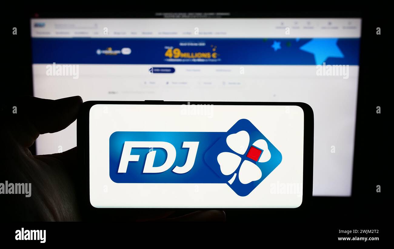 Person holding cellphone with logo of French lottery company Francaise des Jeux (FDJ) in front of business webpage. Focus on phone display. Stock Photo