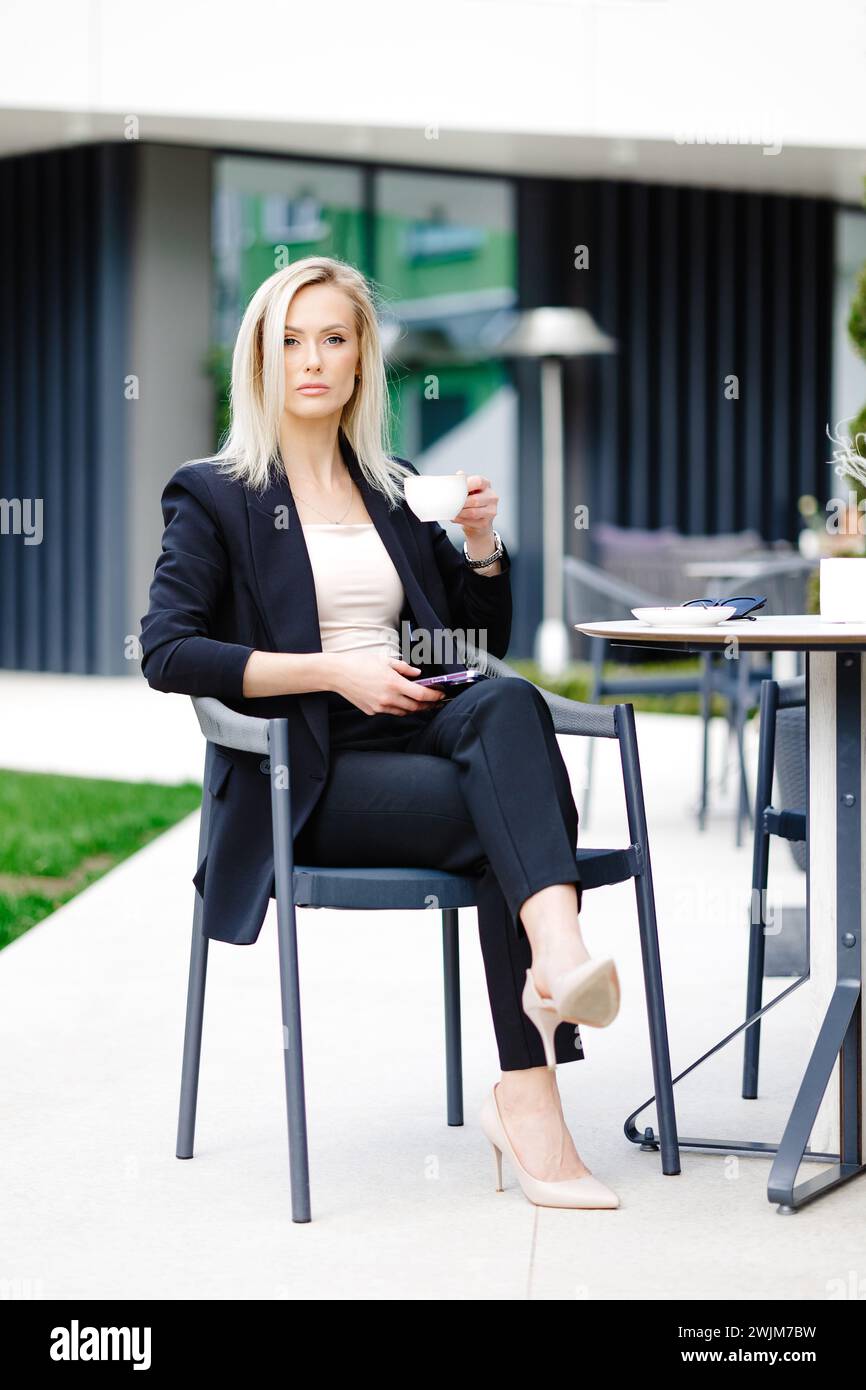 Blonde business lady looks at the phone, reading messages. Woman is relaxing in a cafe on the terrace enjoyment of life. Stock Photo