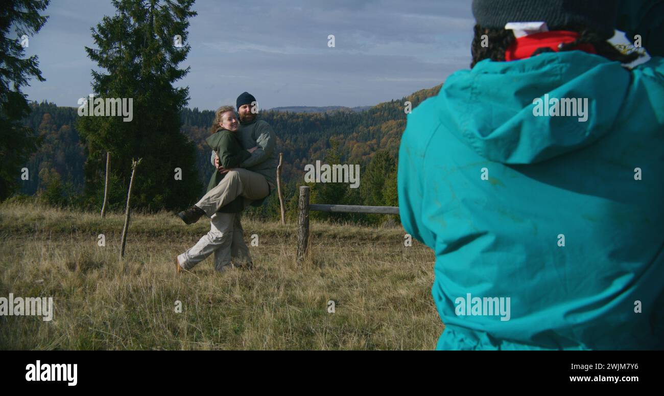 Young hiker takes pictures of Caucasian tourist family using camera. Happy couple of travelers posing on top of hill against beautiful landscape during hiking trip in mountains. Outdoor enthusiasts. Stock Photo