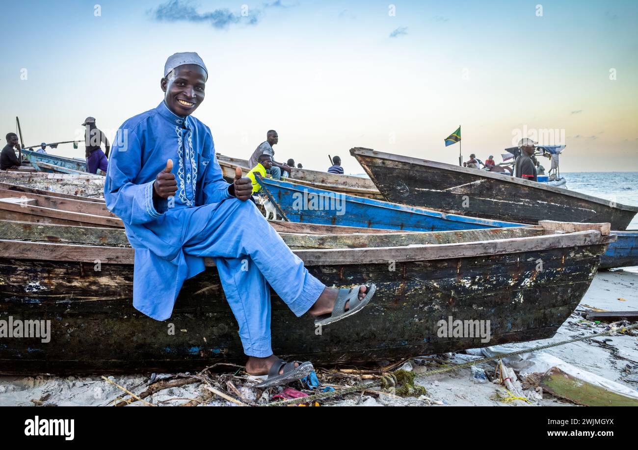 A man wearing a blue kanzu and white embroidered kofia smiles as he sits on a wooden dhow fishing boat at Kivukoni Fish Market in Dar es Salaam, Tanza Stock Photo