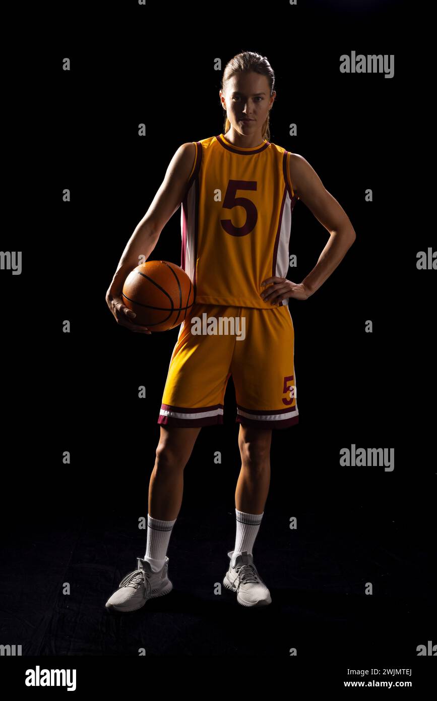 Young Caucasian female basketball player poses confidently in a basketball uniform on a black