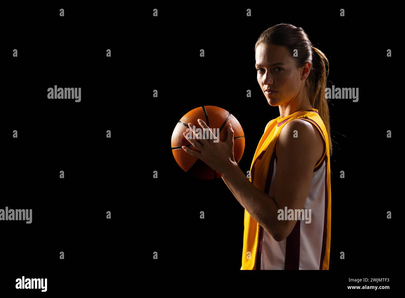 Focused female basketball player shines in a dark setting, exuding confidence. Stock Photo