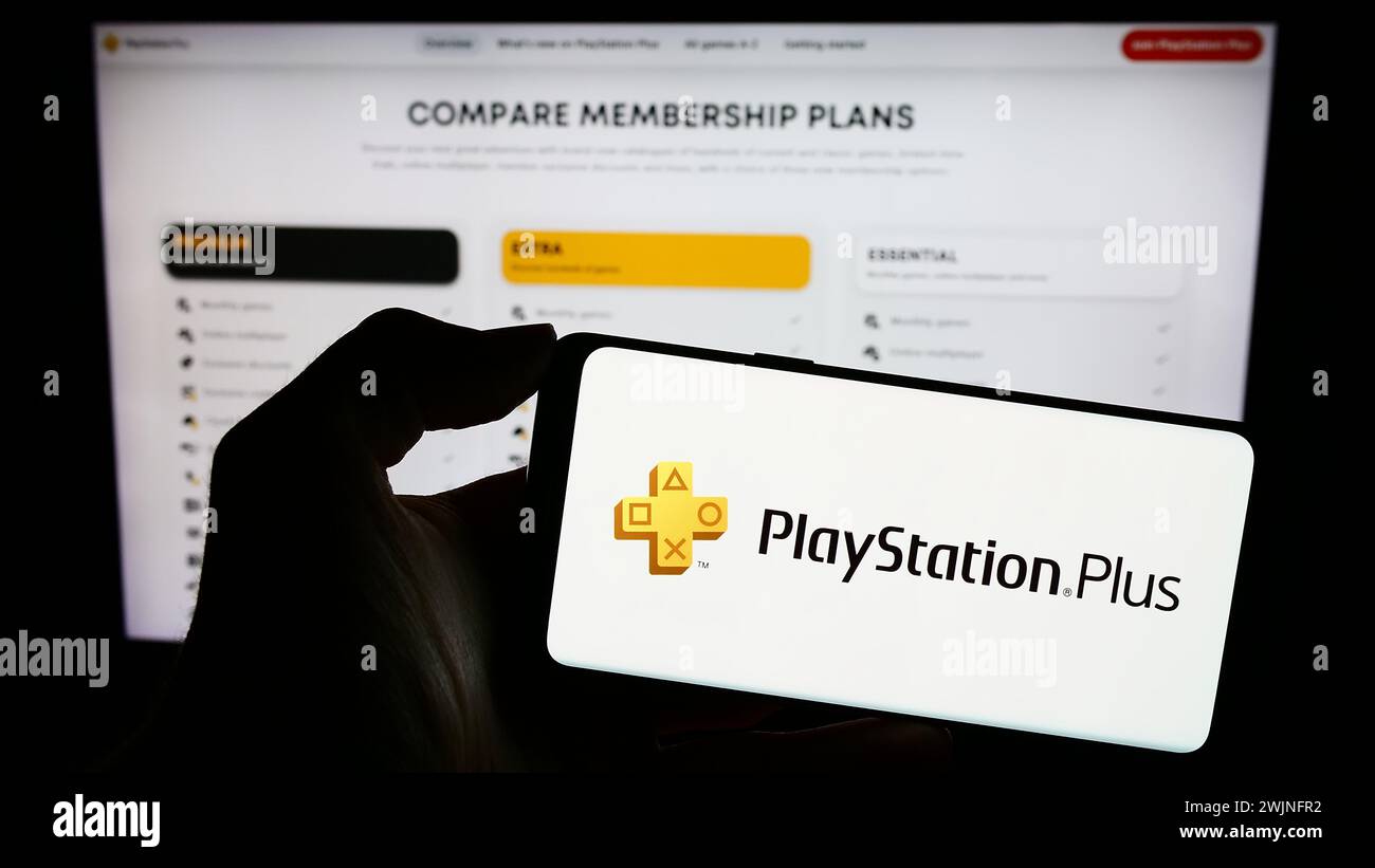Person holding mobile phone with logo of subscription service PlayStation Plus (PS Plus) in front of business web page. Focus on phone display. Stock Photo