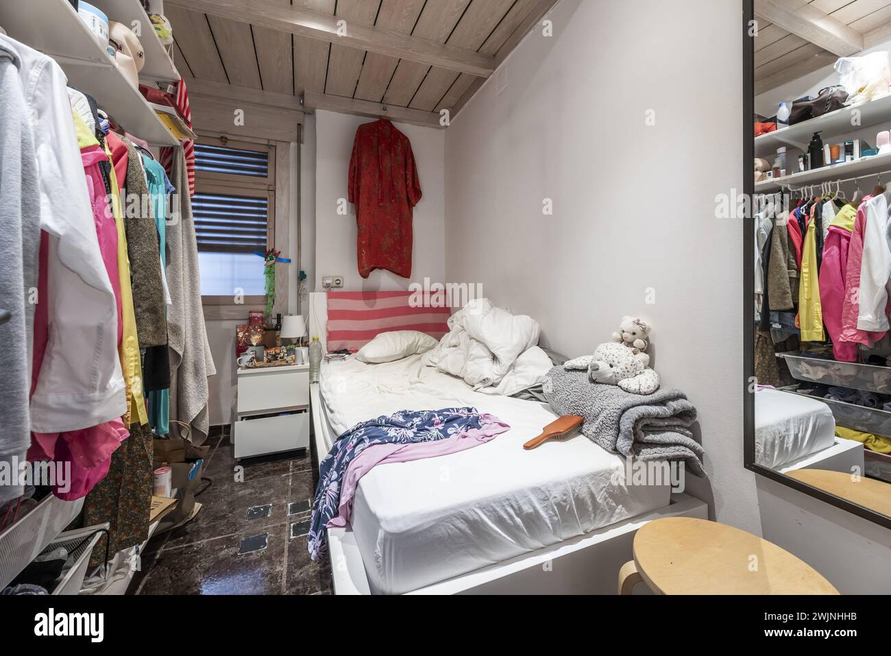 Very small bedroom with a single bed with a large mess of clothes on the mattress, clothes hanging on a hanger and ceilings with tongue and groove woo Stock Photo