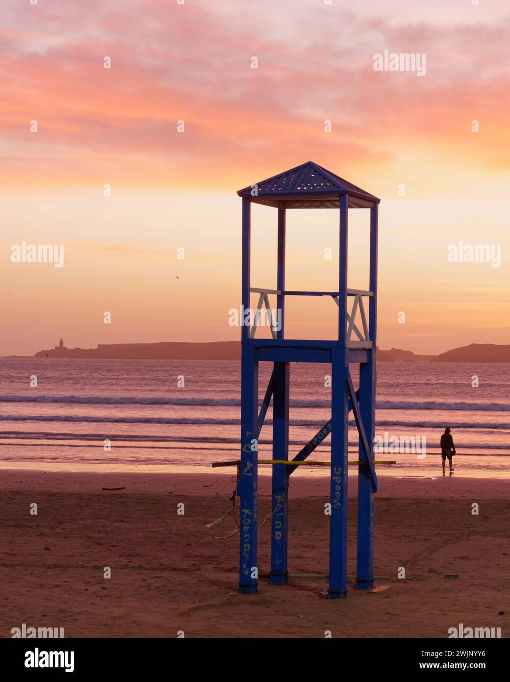 Man in silhouette beside a Wooden lifeguard lookout tower with the sea and an Island behind at sunset in Essaouira, Morocco. February 16th, 2024 Stock Photo