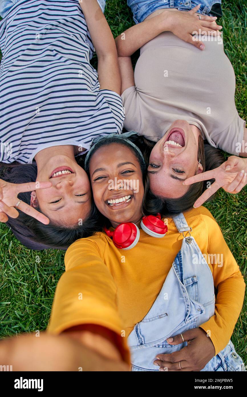 Vertical selfie above three cheerful multiracial young women lying on grass outdoors. Gen z in park. Stock Photo