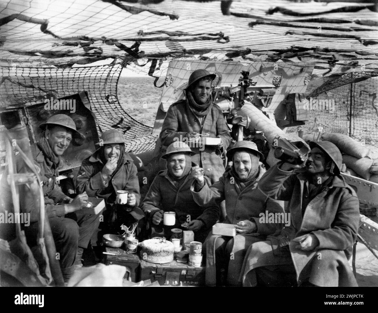 A gun crew having their Christmas dinner complete ***** Christmas cake. Tin Hats and Balaclava helmets were dress for the day when these Australian gunners outside Bardia drank up their Christmas dinner in 1942. The ammunition box table is crowned by a cake from home. December 25, 1940. Stock Photo