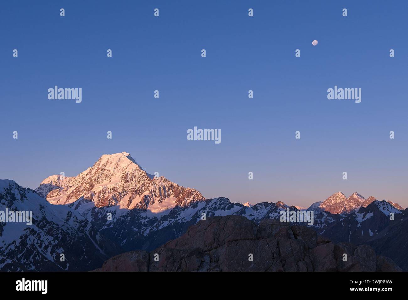 Scenic Sunrise at Mount Cook: Snowy Mountain Peaks Glowing in Golden Light, Accompanied by the Moon in New Zealand's Breathtaking Landscape Stock Photo