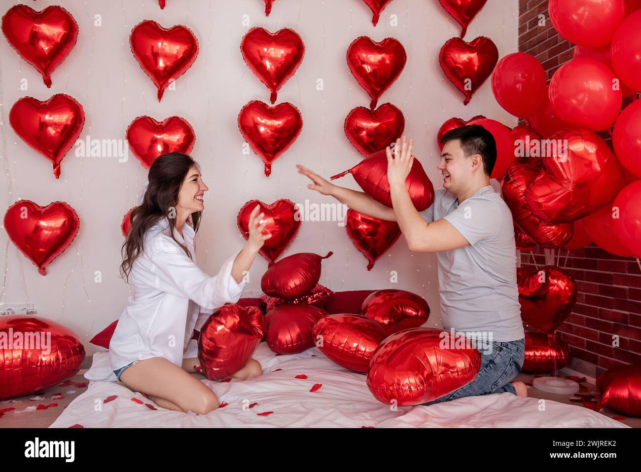 Playful moment between couple engaging in pillow balloons fight on bed, with heart shaped red balloons in background. Woman is joyfully fooling around Stock Photo