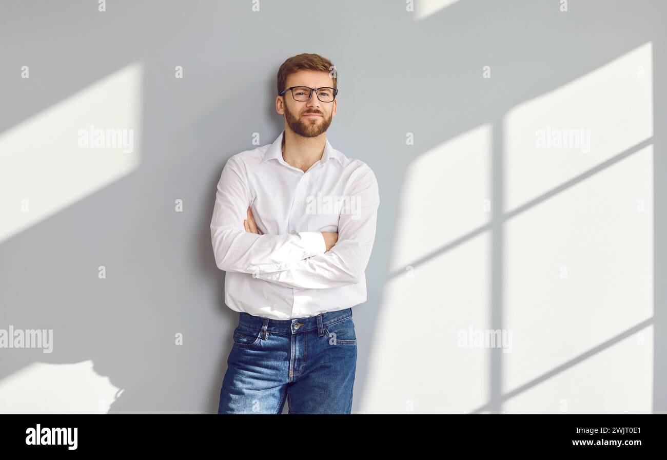Confident bearded man in eyeglasses posing with crossed arms at gray wall Stock Photo