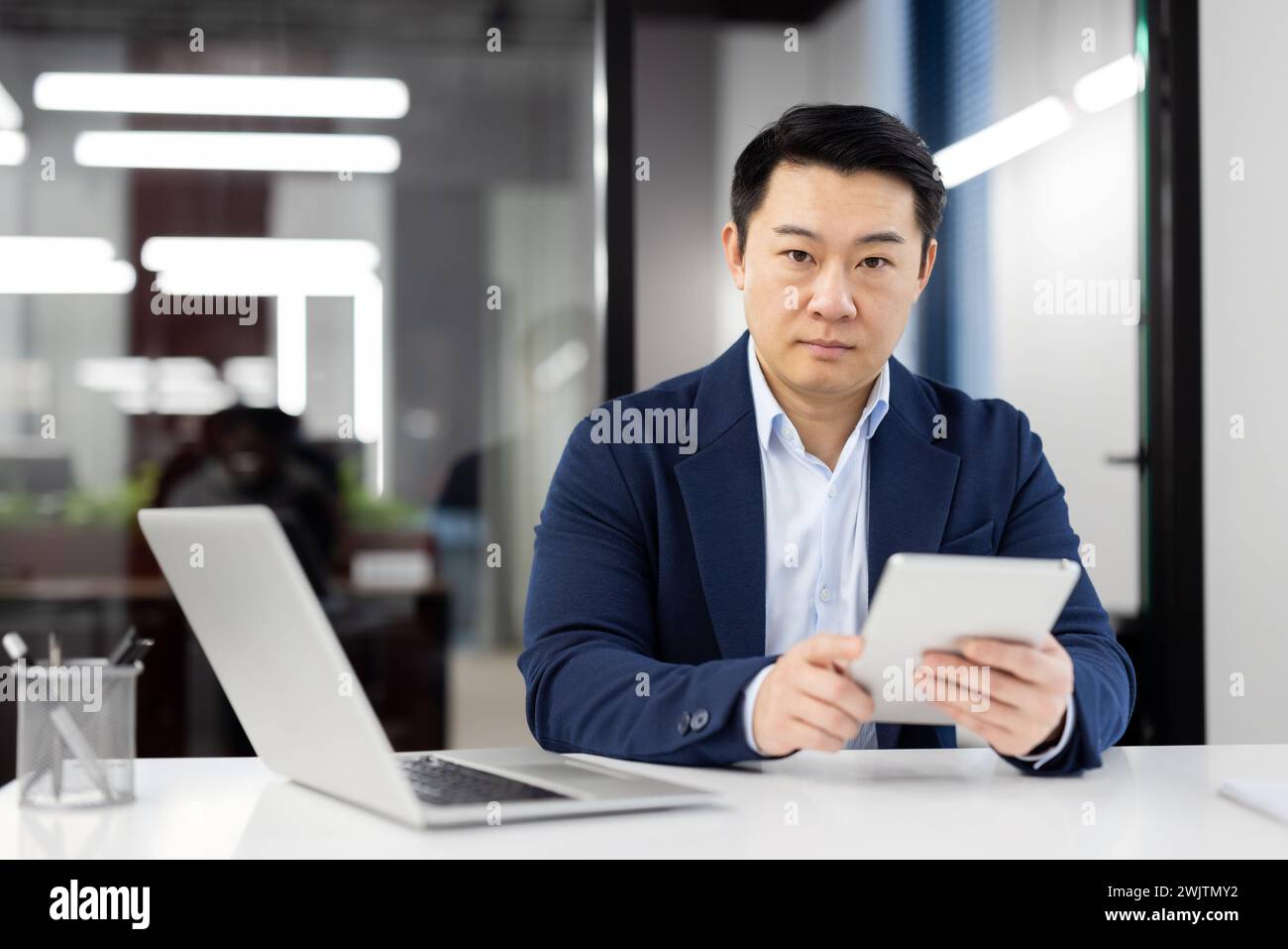 Focused professional businessman in a suit using a tablet at his desk with a laptop in a contemporary office setting. Stock Photo