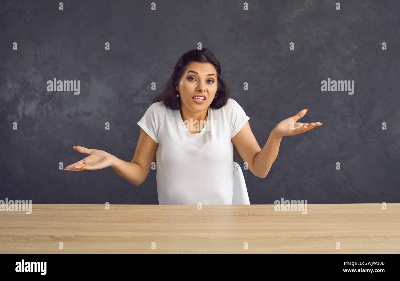 Young caucasian unsure woman sitting at a table shrugs saying that she can't help you. Stock Photo