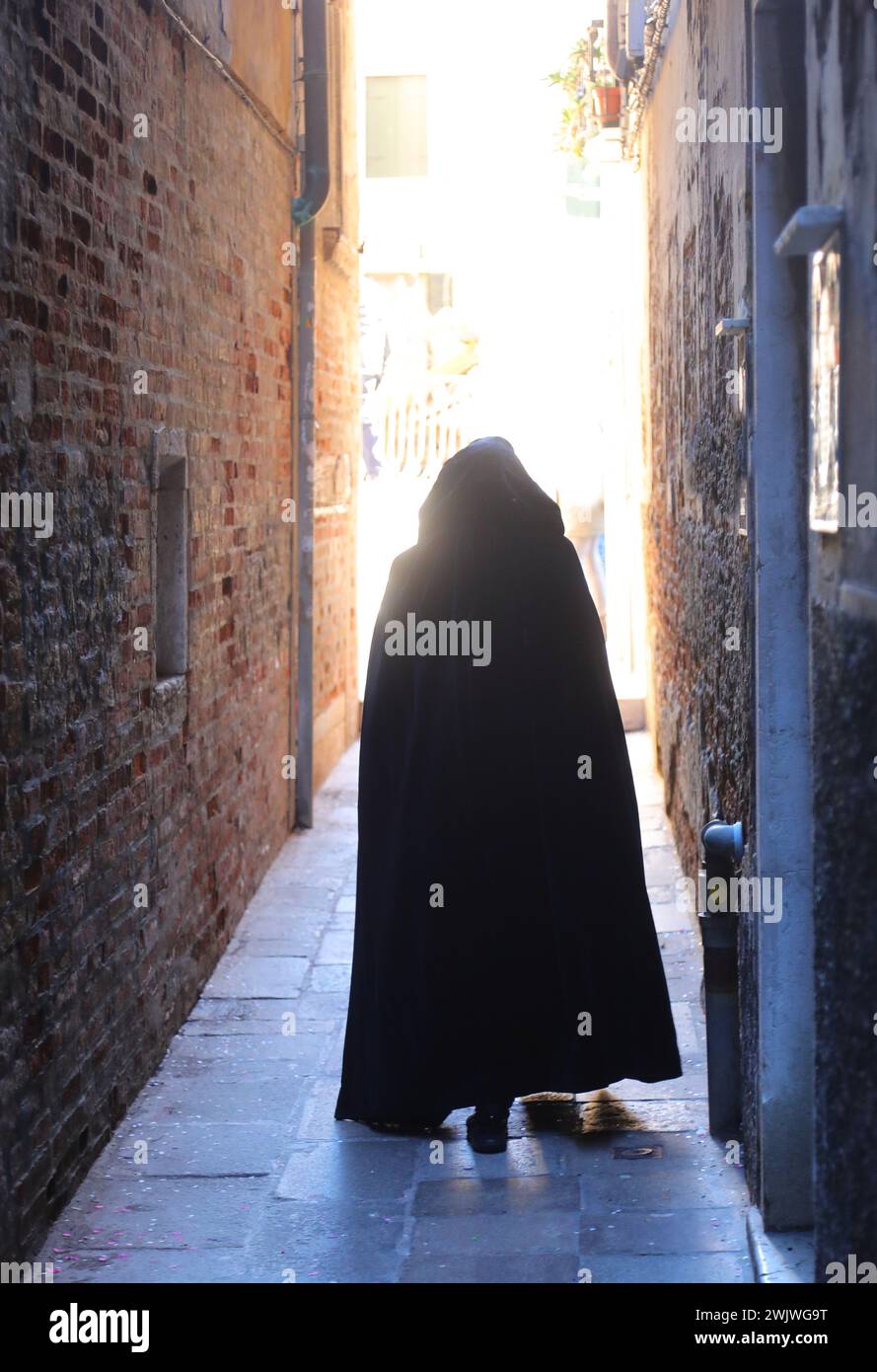 anonymous hooded stroller with black cloak dress walking through the narrow alleys of Venice Stock Photo