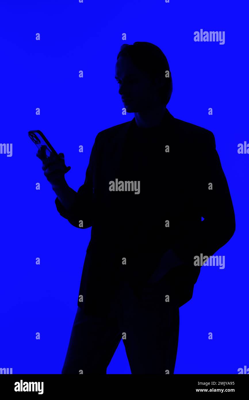 Silhouette an elegant businessman using a modern smartphone in a jacket standing on a blue glowing background, a futuristic concept Stock Photo