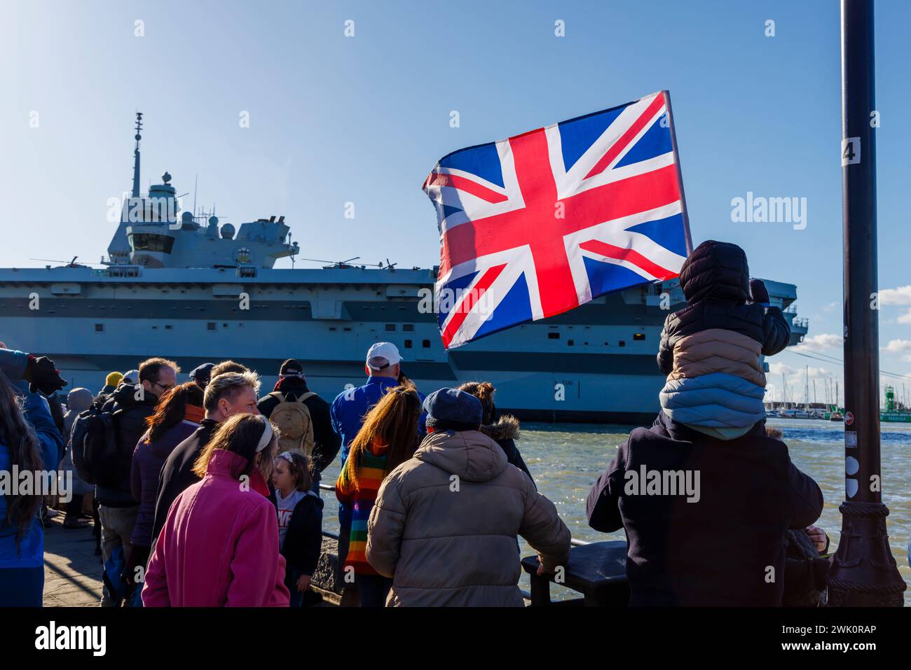 A boy sitting on his father's shoulders waves a Union Jack as Queen Elizabeth-class aircraft carrier 'HMS Prince of Wales' departs Portsmouth Harbour Stock Photo