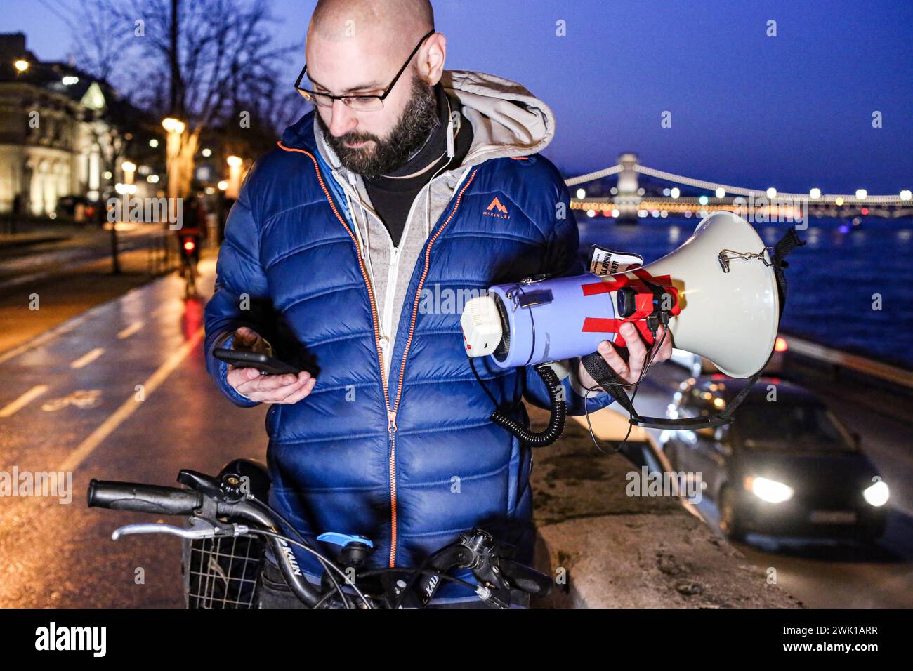 Budapest, Budapest, Hungary. 17th Feb, 2024. Hungarian activist, Balasz T., holds a bull horn and looks at his phone with the chain bridge in back ground along the Danube after a speech where Hungarian Prime Minister VIKTOR ORBAN made his annual speech and comments on three resignations of Hungarian President, KATALIN NOVAK; former Minister and leadership of Hungarian Reform Church, ZOLTAN BALOG and former Justice Minister, JUDIT VARGA following exposure of a man pardoned last year from covering up pedophilia in a children's home. The reveal has caused embarrassment and tarnished the imag Stock Photo