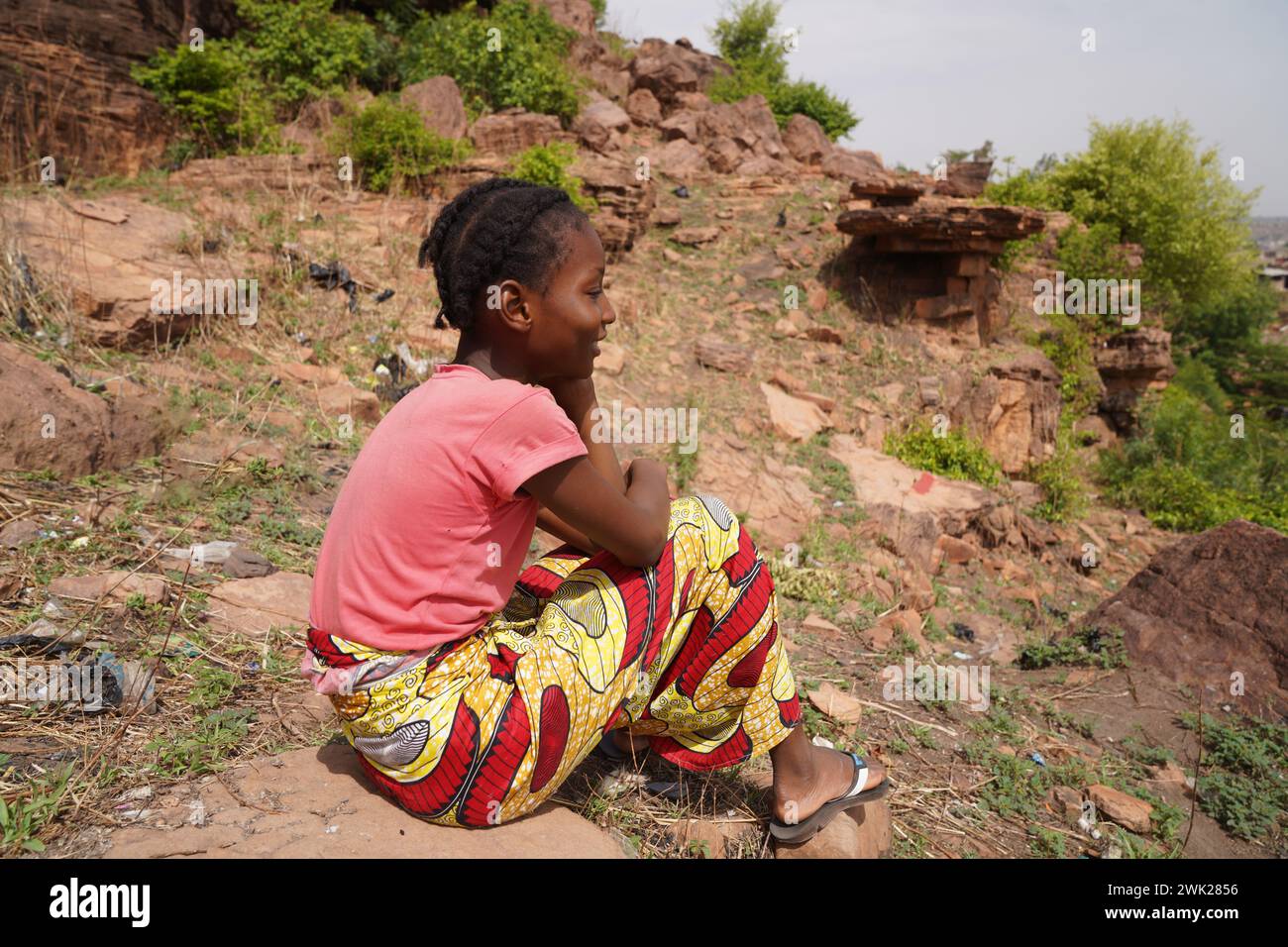 Side view of a serene daydreaming African villagegirl sitting on a rock amidst weeds and brushwood on a sunny day symbolizing differences in lifestyle Stock Photo