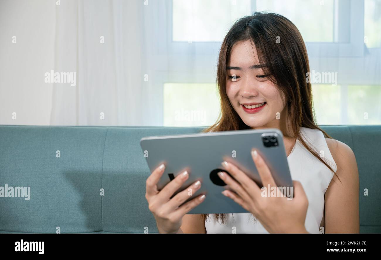 Happy Asian woman playing smart tablet at home while relaxing on sofa ...