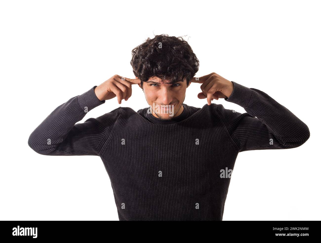 Too much noise: attractive young man covering his ears. A young man with curly hair is seen wearing a black sweater. He is standing against a neutral background, with the focus on his clothing and hair. Stock Photo