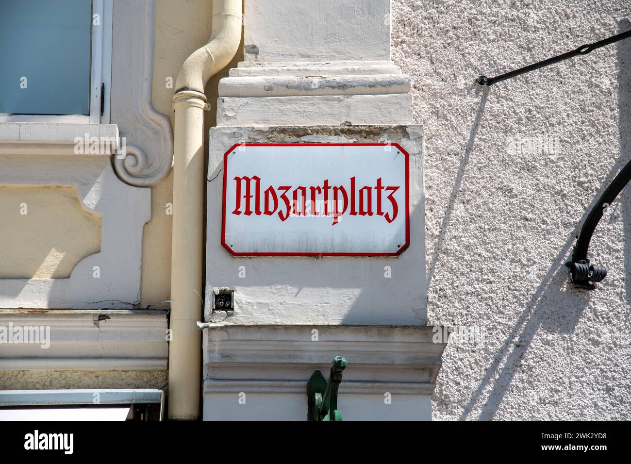 A white metal sign with the inscription Mozartplatz in old German lettering. The sign hangs on a house wall and has red letters and a red border. Stock Photo