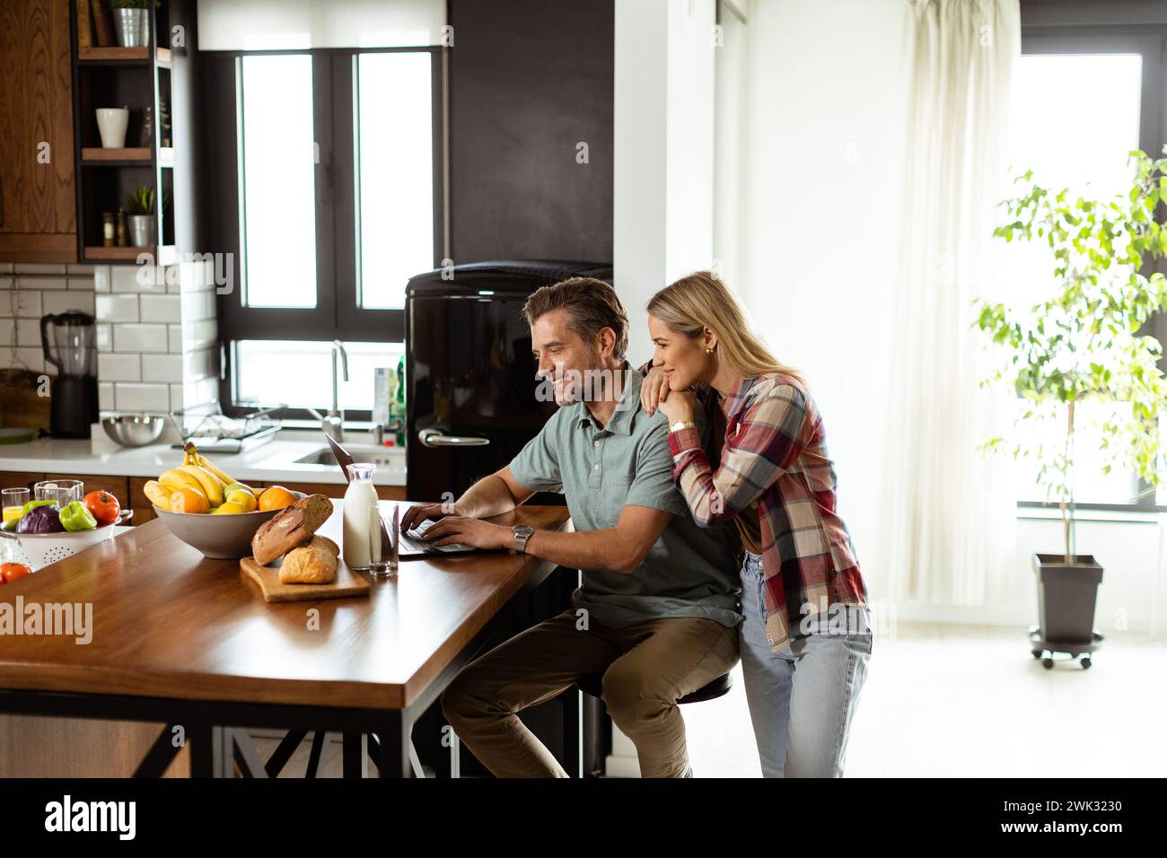 Smiling couple shares a moment of togetherness as they sit by the kitchen counter, surrounded by a spread of fresh fruits and pastries, bathed in the Stock Photo