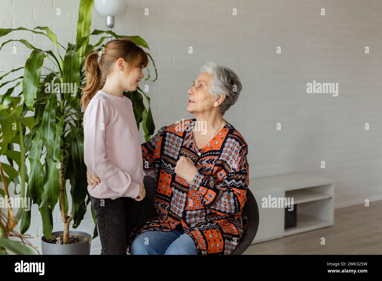 In a softly-lit room, a young girl stands beside a potted plant, sharing a moment of connection and conversation with her elderly grandmother, the joy Stock Photo