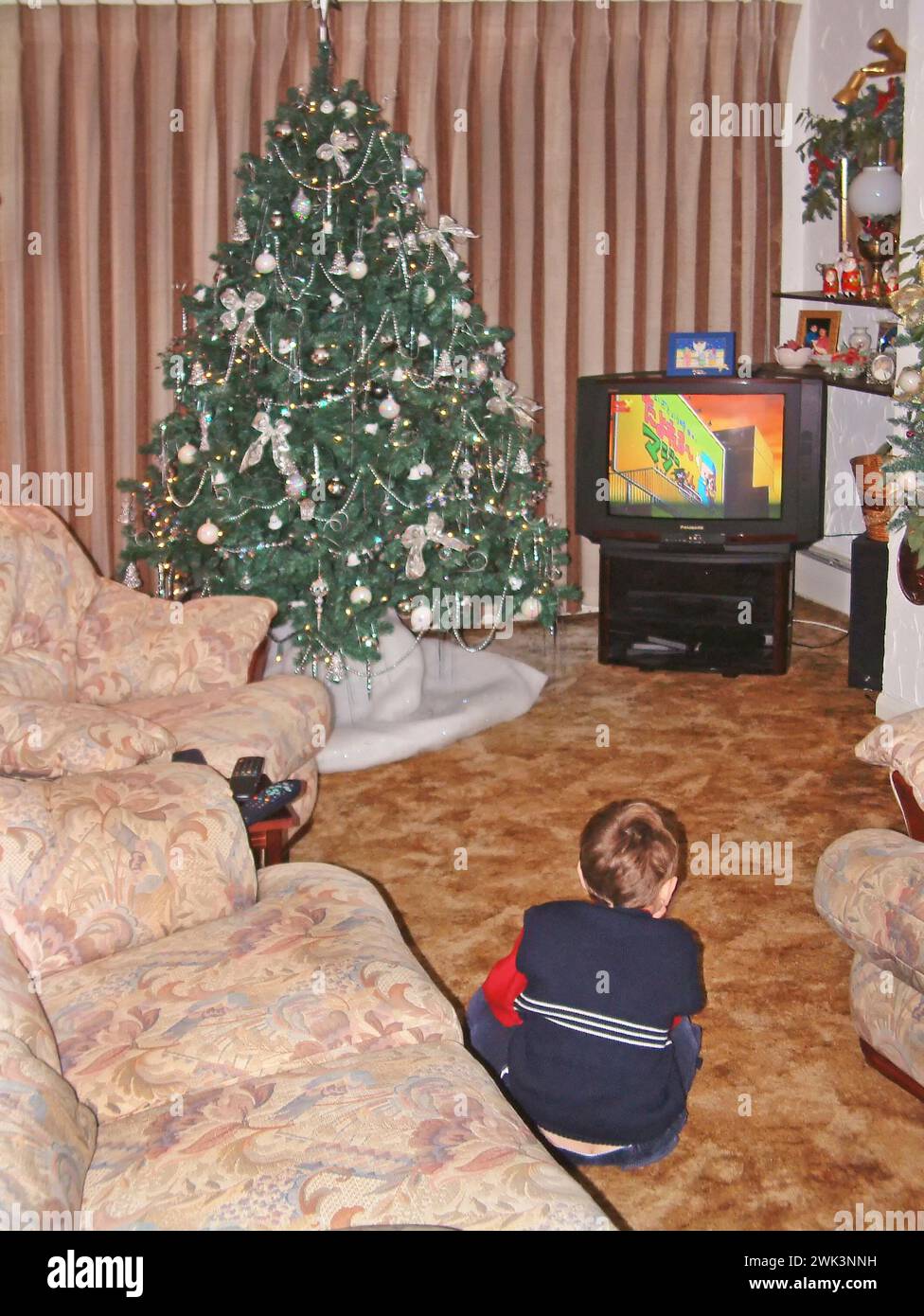 1990s three year old boy historical archive back view image watching TV old fashioned coloured television screen beside decorated Christmas tree sitting on carpet in 90s furnished house living room with curtains drawn Essex England UK Stock Photo