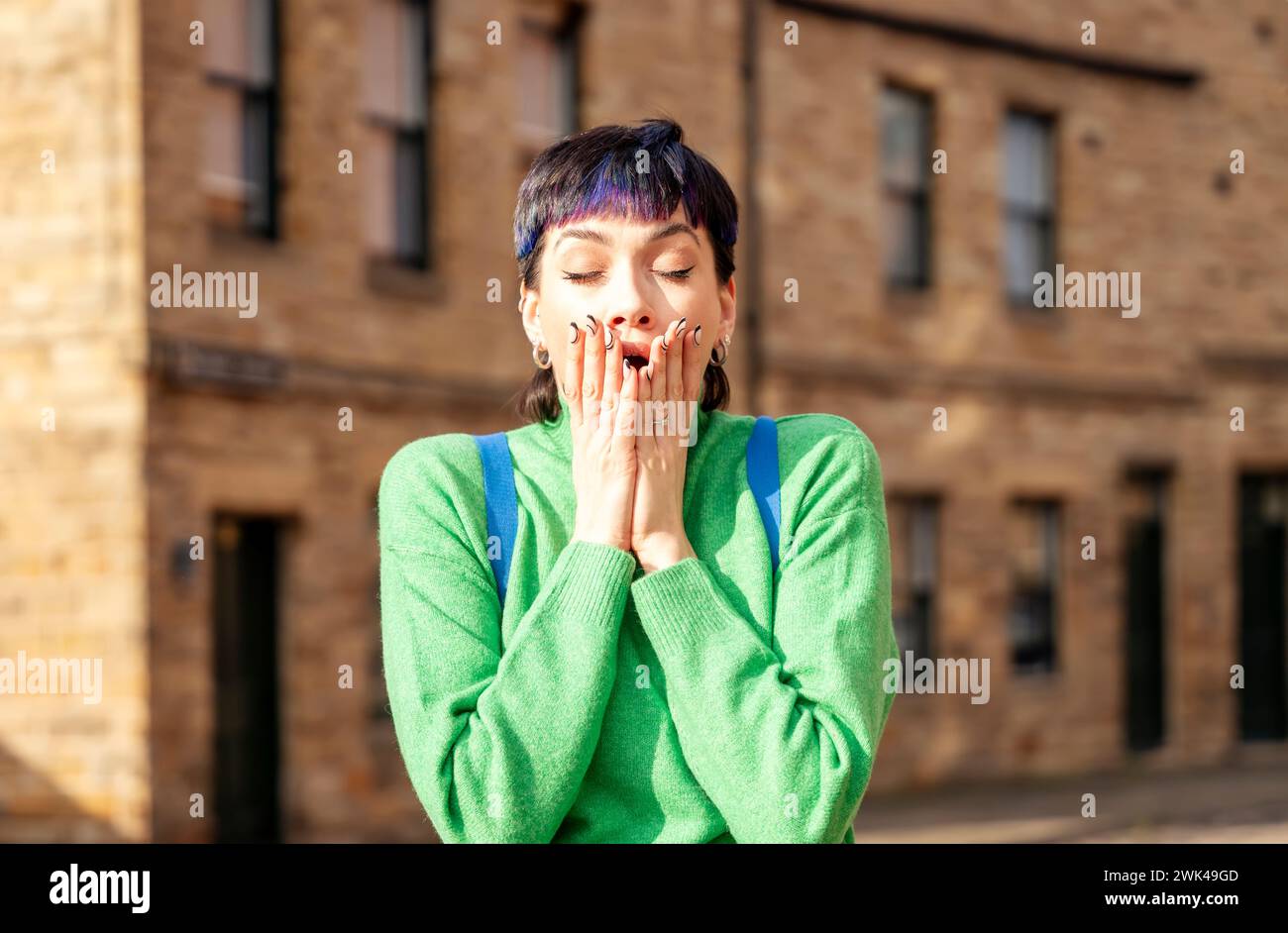 a woman in a green shirt shocked from news.  Holding hands on her mouth. Standing alone outside. Old buildings behind. Stock Photo