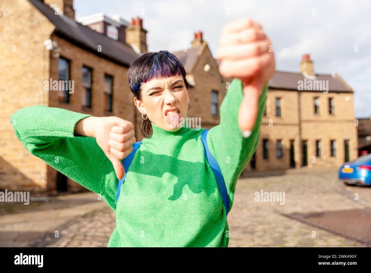 woman in green sweater giving thumb down gesture, dislikes something, having disgusting expression, showing disapproval sign outside and tongue. Negat Stock Photo