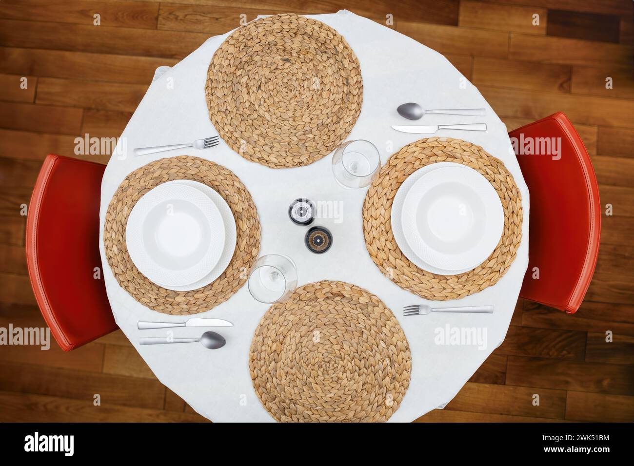 Top view of a round dining table set with white plates, silverware, woven placemats, and red chairs, ready for a meal. Stock Photo