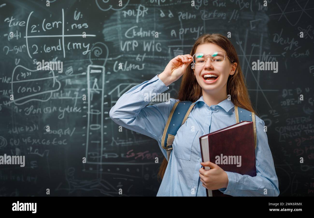 Back to school. Happy cute industrious teenager indoors. Girl is learning in class on background of blackboard. Stock Photo