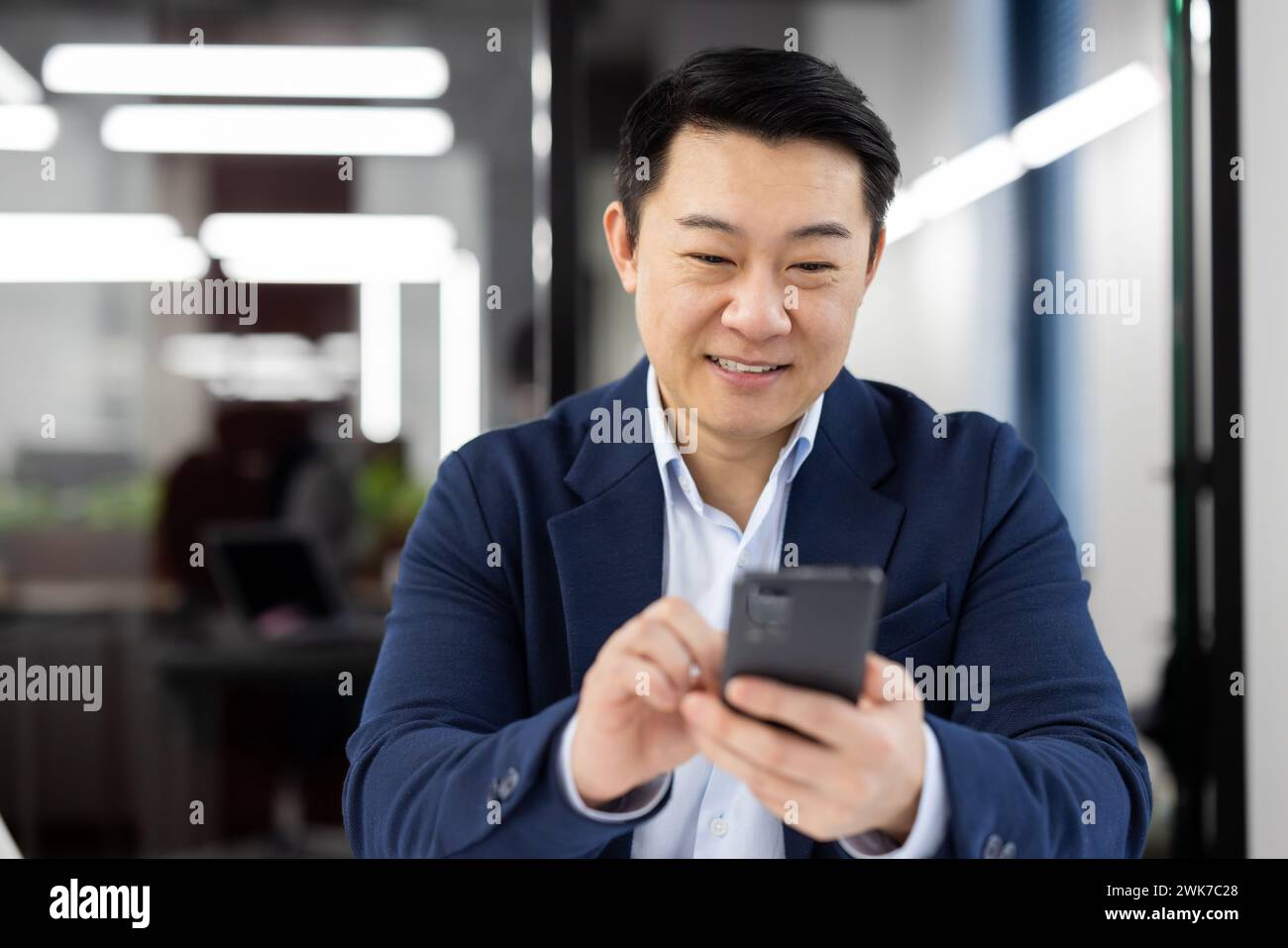 Asian man inside office with phone in hands, smiling contentedly ...
