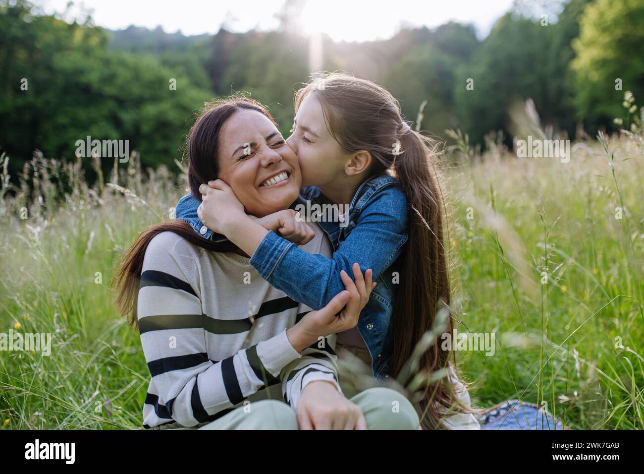 Beautiful mother with daughter, embracing or hugging, sitting in the grass at meadow. Concept of Mother's Day and maternal love. Stock Photo