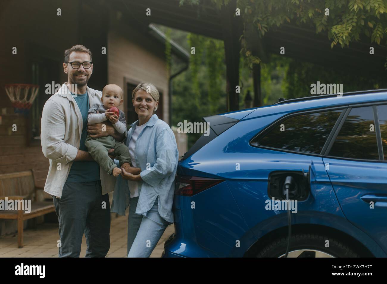 Young family with little baby standing by their electric car. Electric vehicle with charger in charging port. Stock Photo