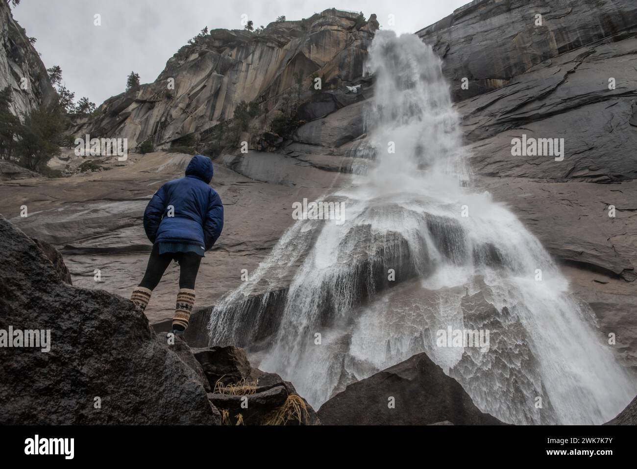 A female hiker stands underneath a cascading waterfall in Yosemite National Park in the Sierra Nevada Mountains of California, USA. Stock Photo