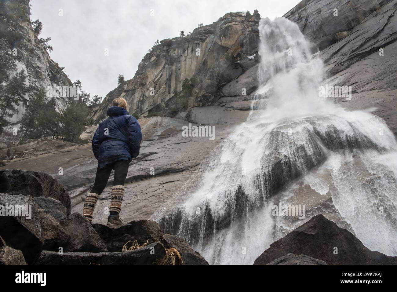 A female hiker stands underneath a cascading waterfall in Yosemite National Park in the Sierra Nevada Mountains of California, USA. Stock Photo