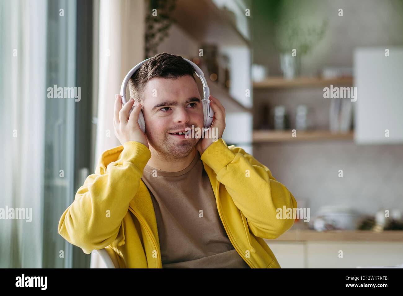 Young man with down syndrome listening his music via headphones. Sitting in the kitchen, wireless headphones on head. Stock Photo